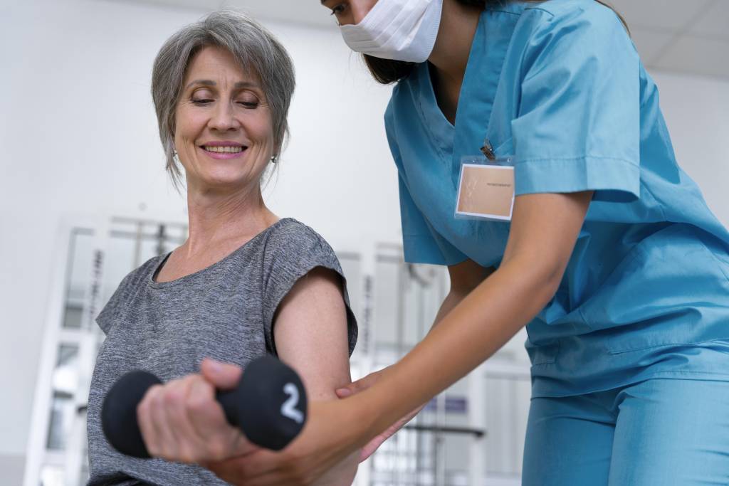 Medical assistant guiding senior woman with physiotherapy exercises, promoting comfort and movement in aged care therapy.